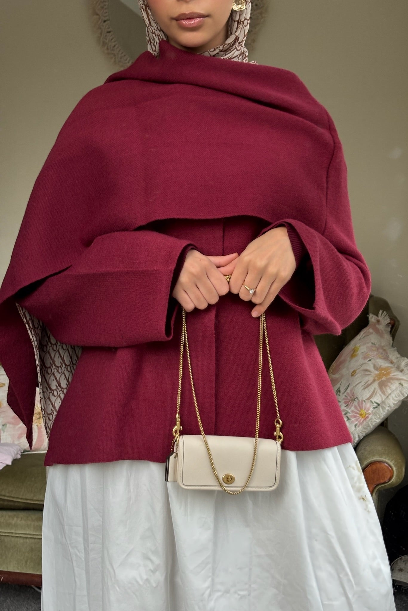 Person wearing a burgundy cape over a white outfit, holding a cream handbag.