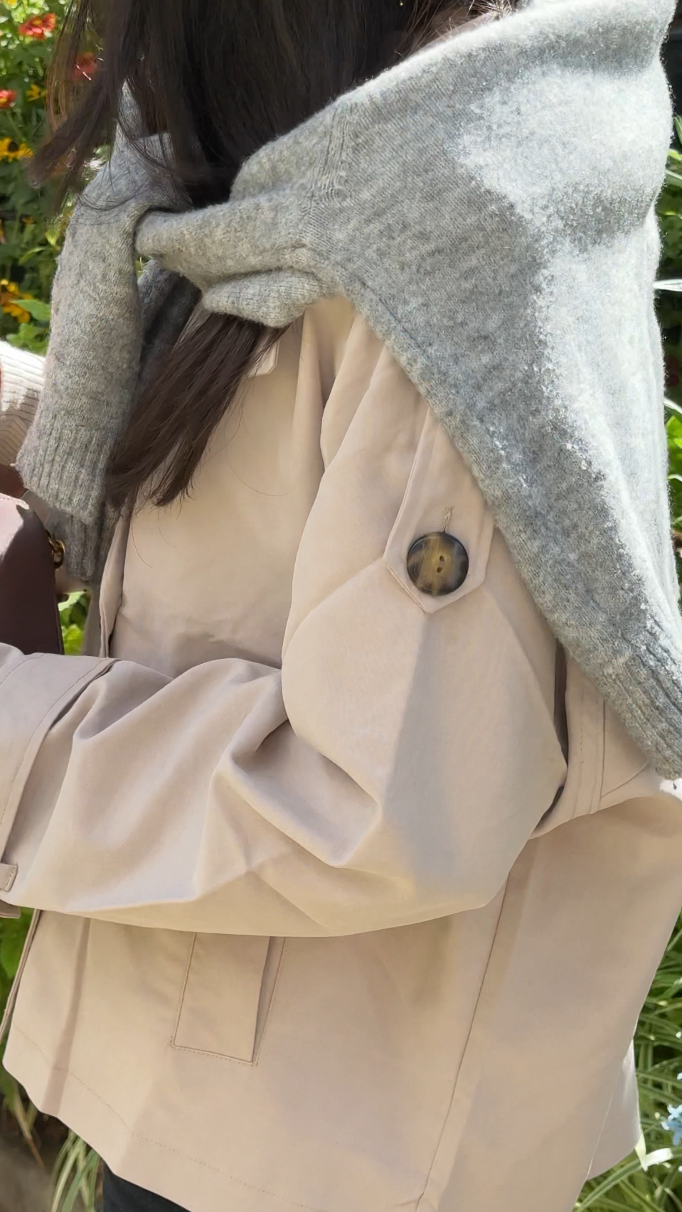 Person wearing a beige jacket with a gray hood in an outdoor setting with greenery and flowers.