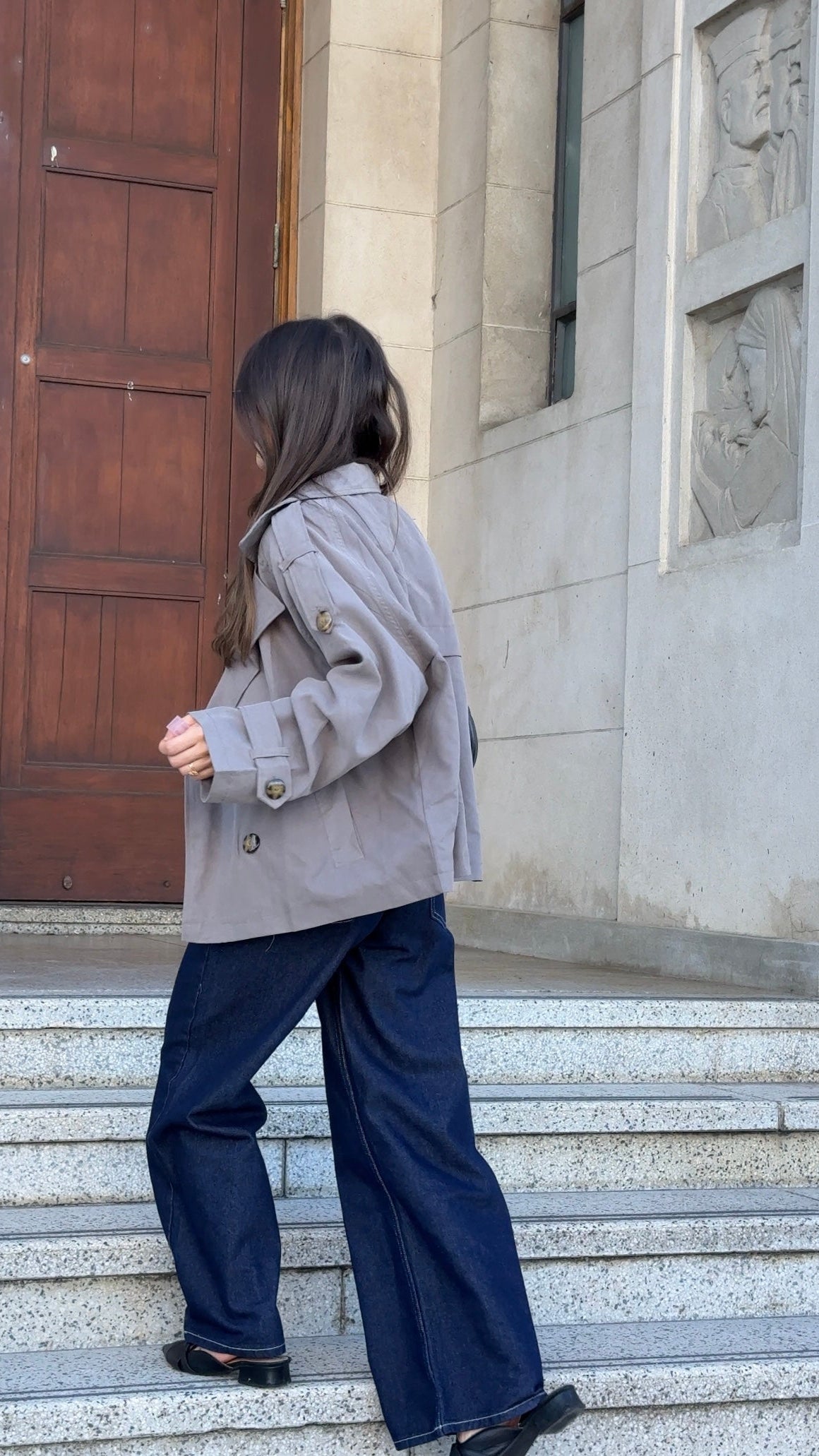 Person wearing a gray coat and blue pants standing on stone steps outside a building.