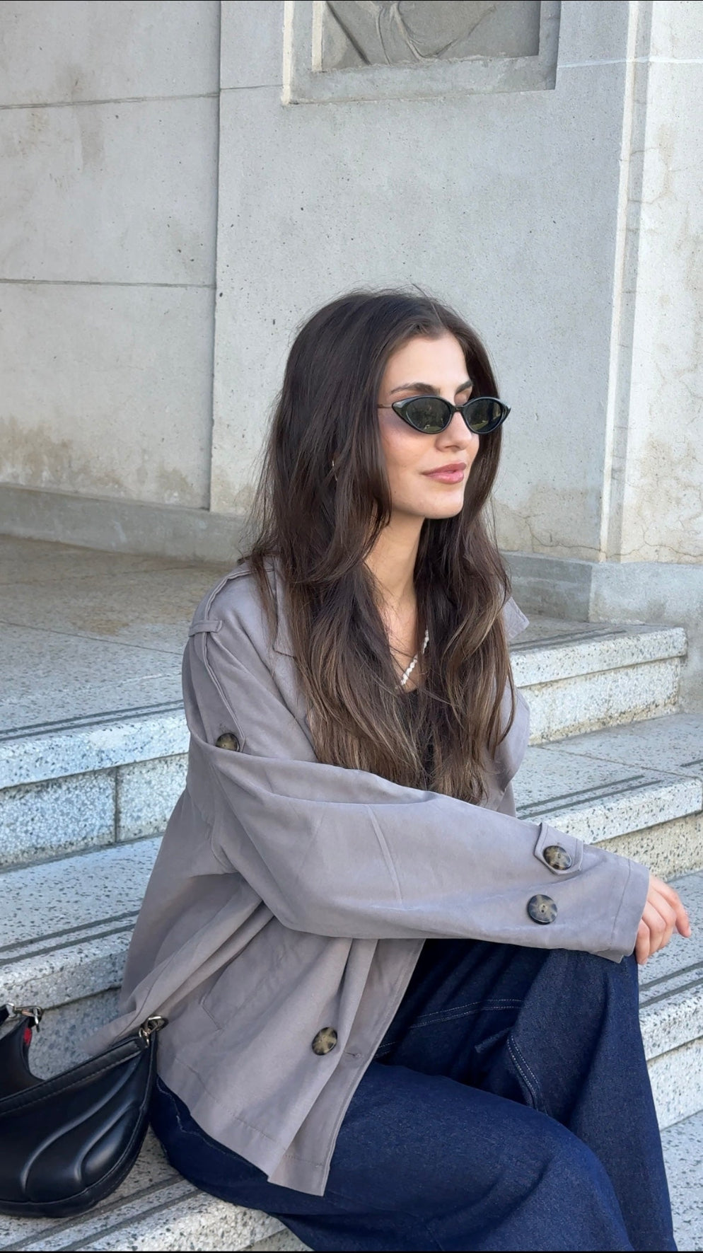 Woman wearing sunglasses and a light gray coat sitting on steps.