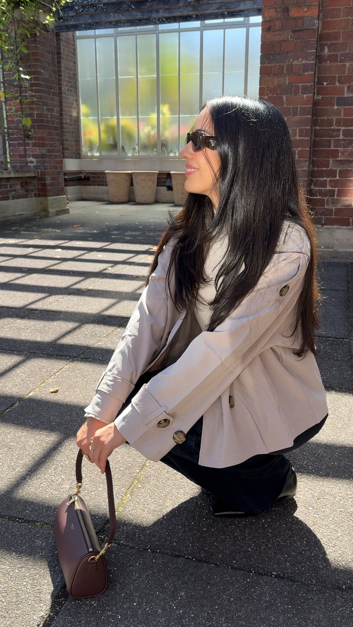 Woman in a beige coat holding a brown handbag on a sunlit pavement.