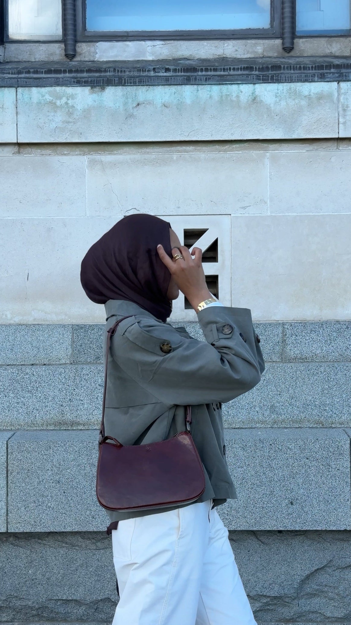 Person wearing a hijab and carrying a maroon handbag against a gray building background