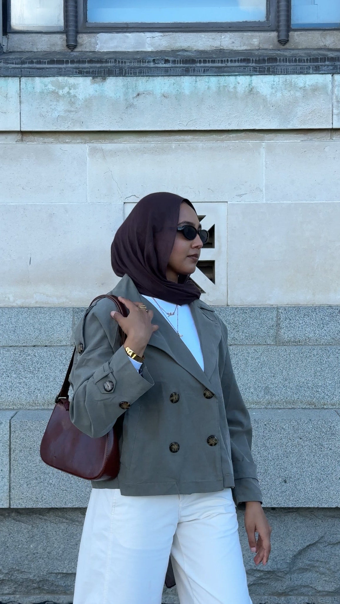 Woman wearing a hijab and gray coat standing against a stone wall.